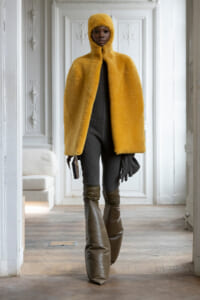 Person wearing a bright yellow fuzzy hooded cape walks forward in a white ornate room, holding a small object in one hand.