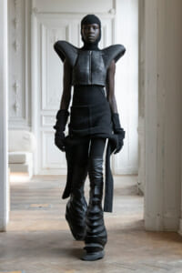 Model in a futuristic black armor-like top with oversized shoulders, balaclava, gloves, and sculpted knee-high boots in a bright ornate room
