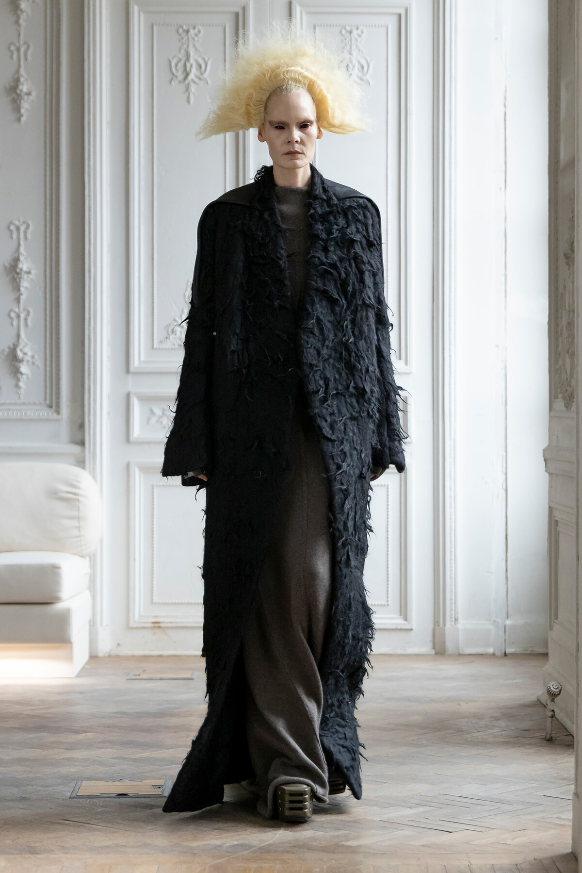 Tall model with dramatic blonde hair wearing a long black textured coat in a white ornate room.