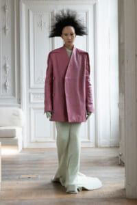 Model in a oversized pink coat with a dramatic black spiky hairstyle, standing in a white ornate room with paneling and molding, wearing light green knit pants.
