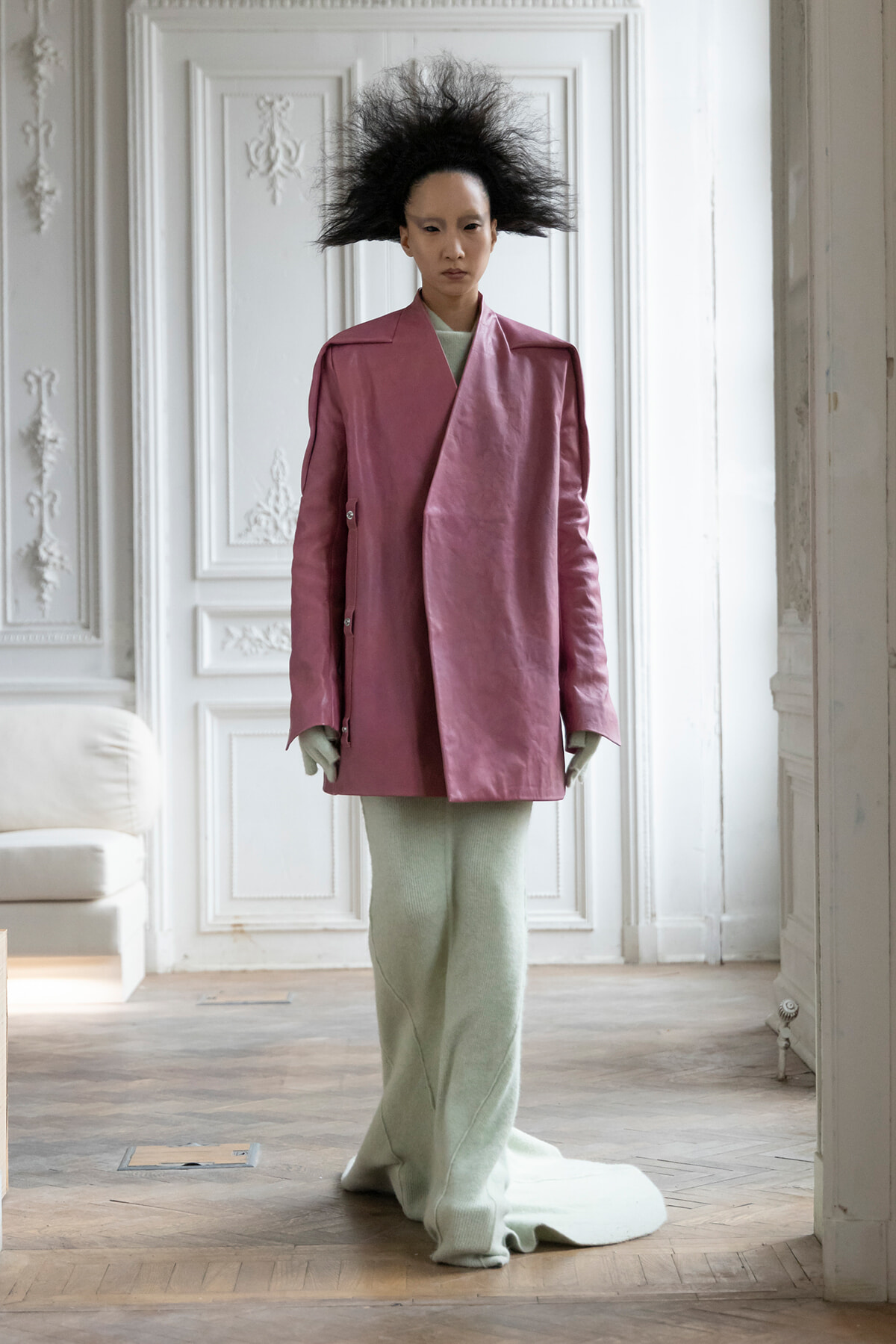 Model in a oversized pink coat with a dramatic black spiky hairstyle, standing in a white ornate room with paneling and molding, wearing light green knit pants.