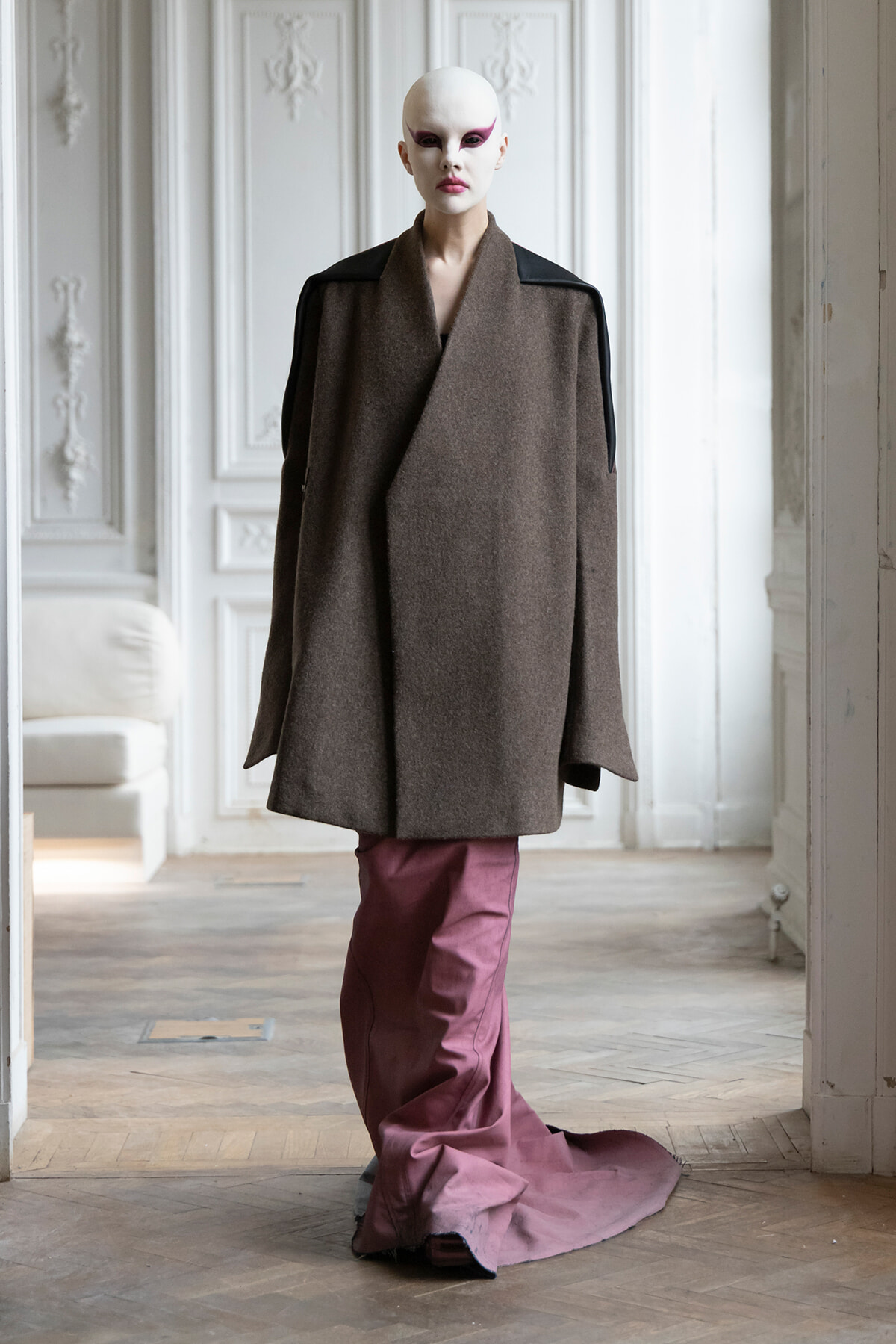 Model with stark white makeup wears an oversized brown wrap coat with black shoulder panels and pink trousers in a pale, ornate room.