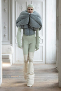 Model wearing an oversized fluffy gray-blue wrap around the shoulders, with a mint knit bodysuit and hat, standing in a white ornate room.