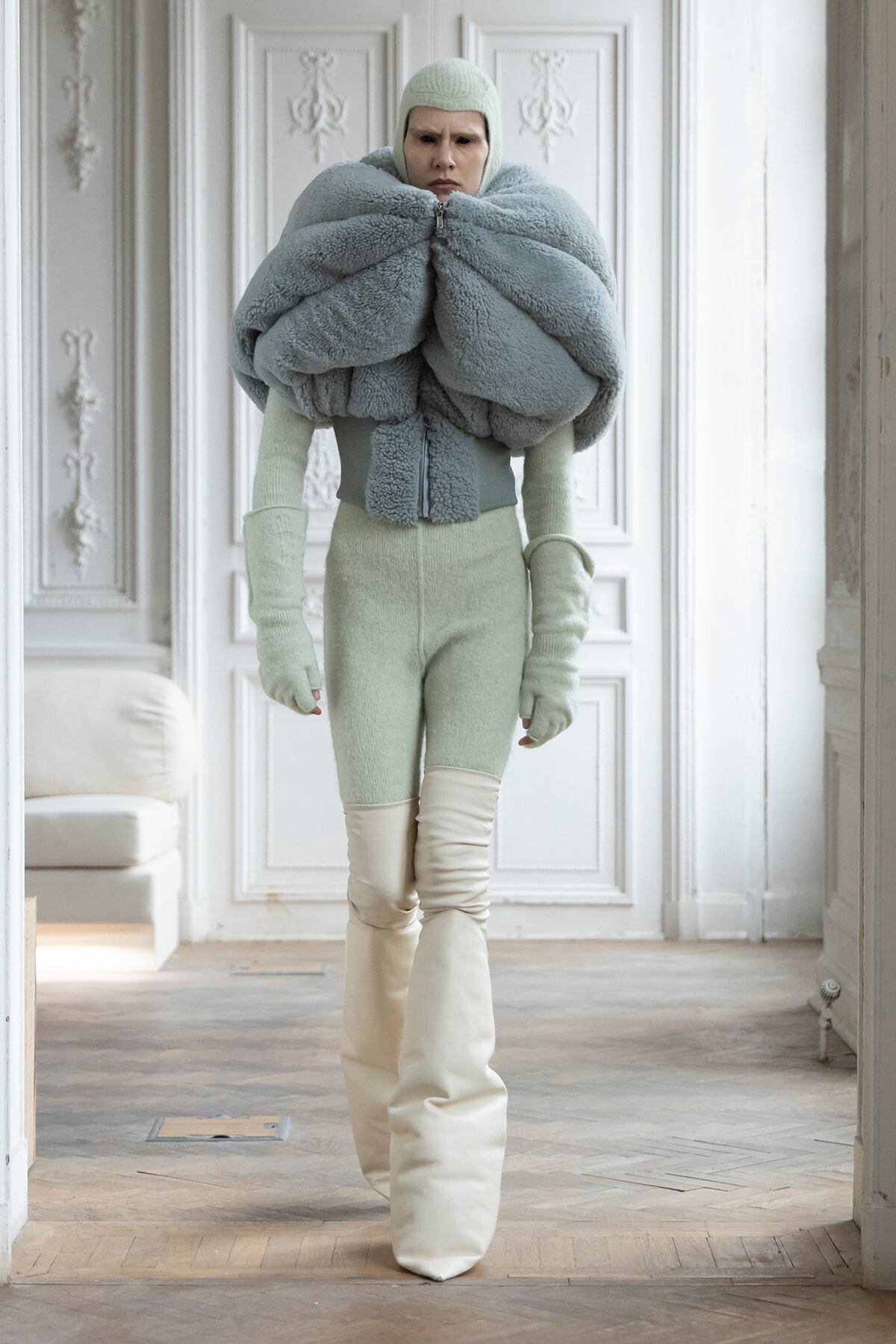 Model wearing an oversized fluffy gray-blue wrap around the shoulders, with a mint knit bodysuit and hat, standing in a white ornate room.