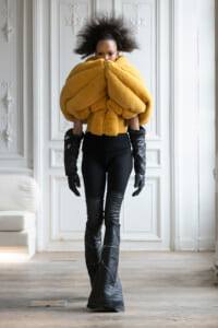 Model in an oversized mustard-yellow fleece jacket zipped up, with black gloves and thigh-high boots in a white, ornate room.