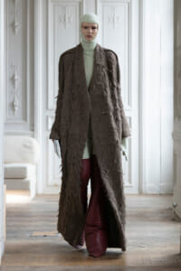 Full-length mannequin wearing a long textured brown coat over a mint knit turtleneck and balaclava, in an ornate white room.