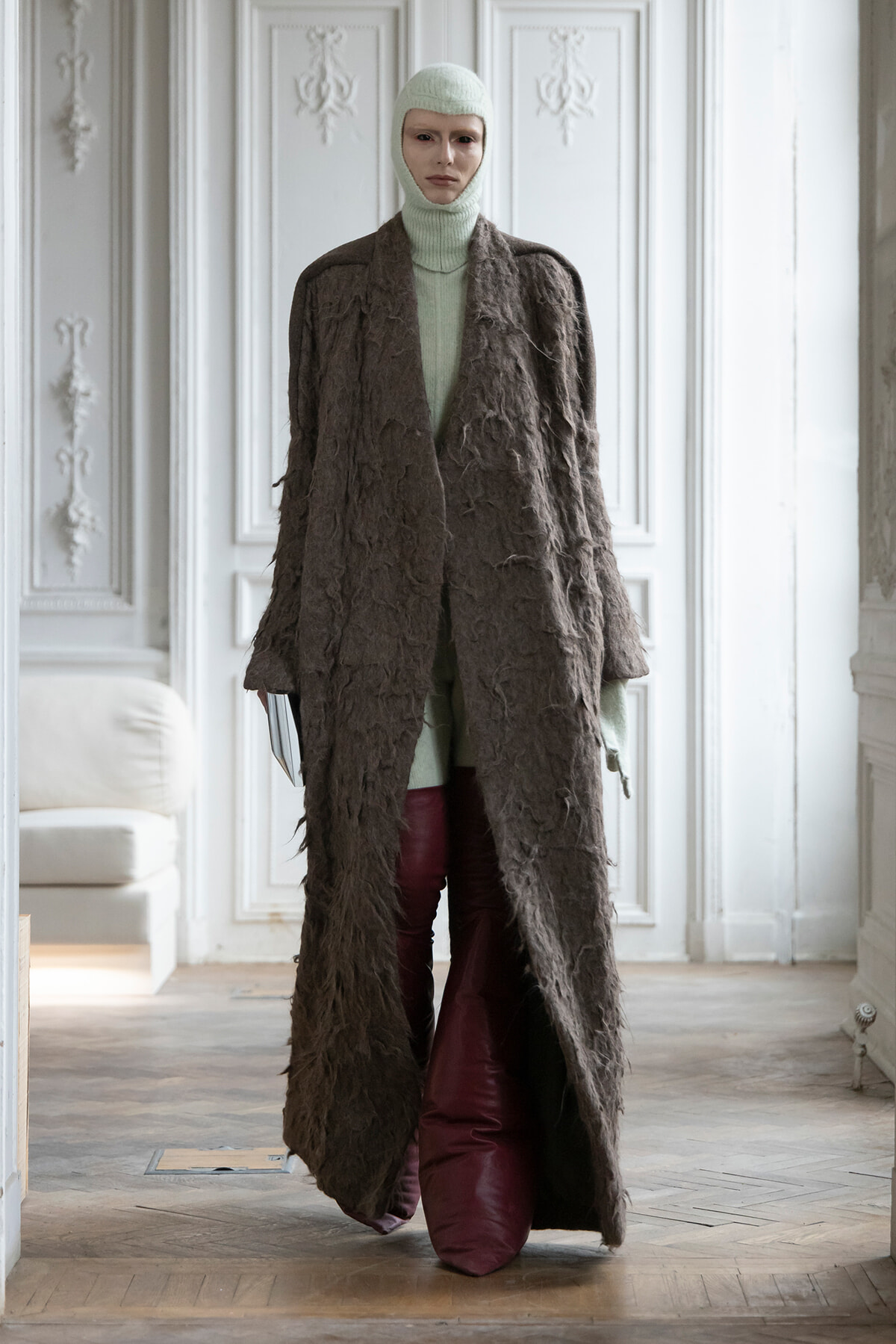 Full-length mannequin wearing a long textured brown coat over a mint knit turtleneck and balaclava, in an ornate white room.