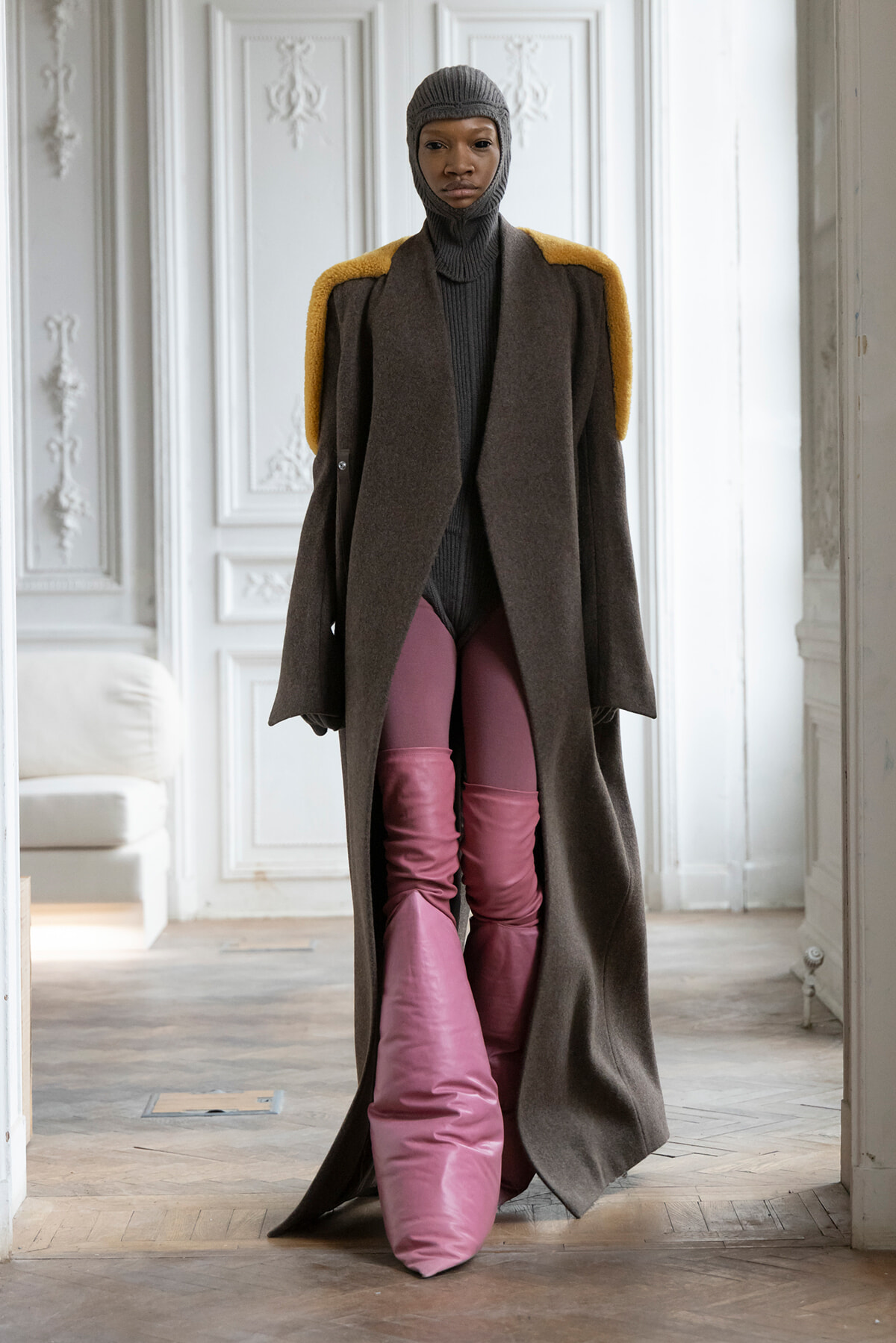Model wearing a long brown coat with yellow shoulder accents, gray knit balaclava, and pink quilted pants in a bright, ornately paneled room.