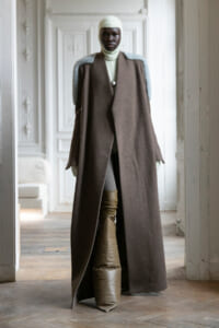 Model standing in an ornate room wearing a long brown coat with pale blue shoulder details, cream knit balaclava and turtleneck.