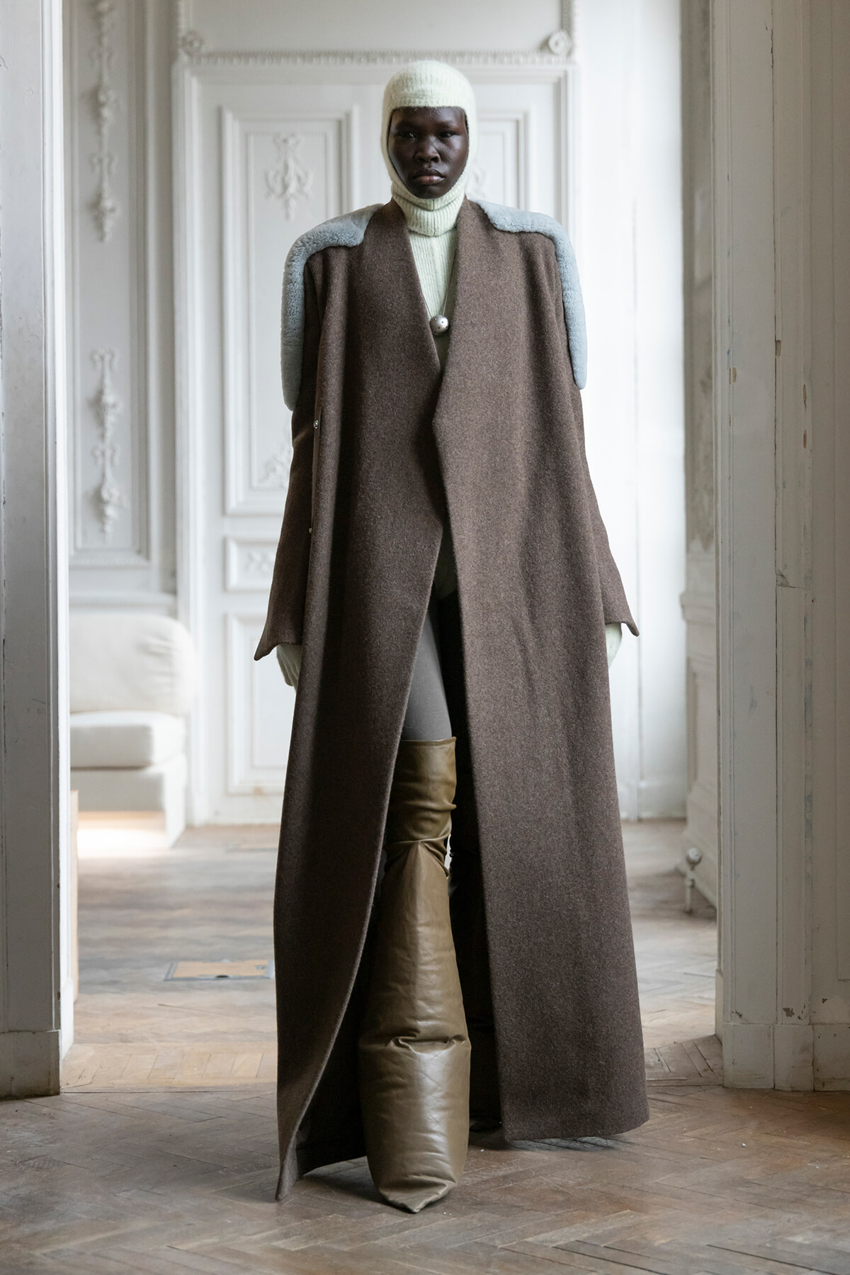 Model standing in an ornate room wearing a long brown coat with pale blue shoulder details, cream knit balaclava and turtleneck.