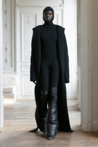 Model in a full black knit outfit (turtleneck, cape-like coat) stands in a white ornate room with detailed mouldings.