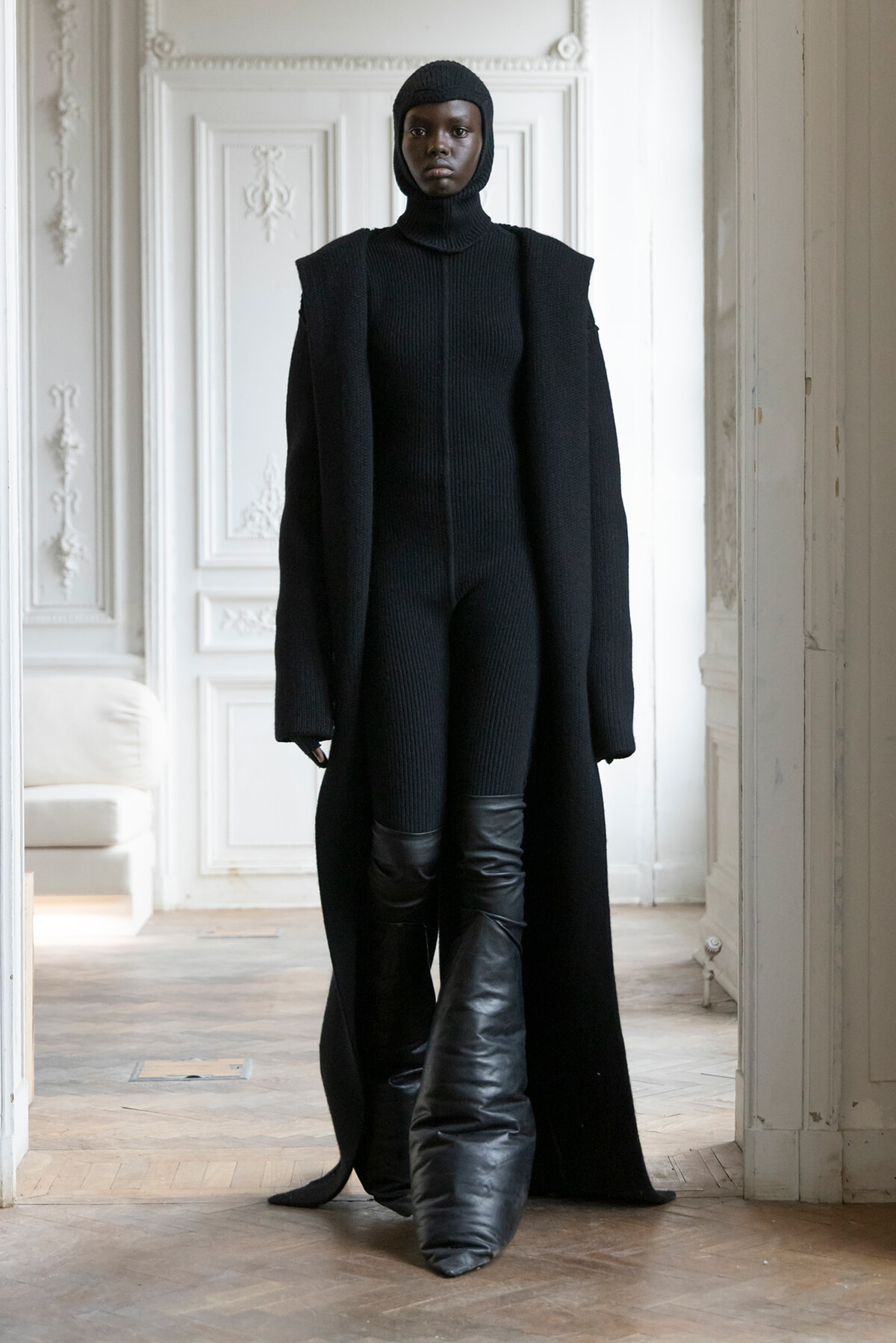 Model in a full black knit outfit (turtleneck, cape-like coat) stands in a white ornate room with detailed mouldings.