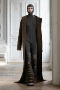 Model wearing a dark ribbed jumpsuit with a balaclava hood and a long brown coat, paired with tall brown strapped boots, standing in an ornate white room.