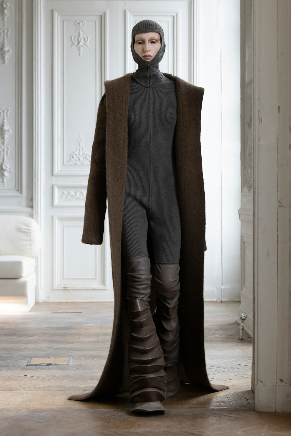 Model wearing a dark ribbed jumpsuit with a balaclava hood and a long brown coat, paired with tall brown strapped boots, standing in an ornate white room.