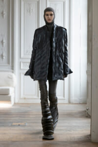 Model wearing an oversized black quilted coat, grey knit balaclava, and dramatic stacked-strap boots in a bright, ornate white room.