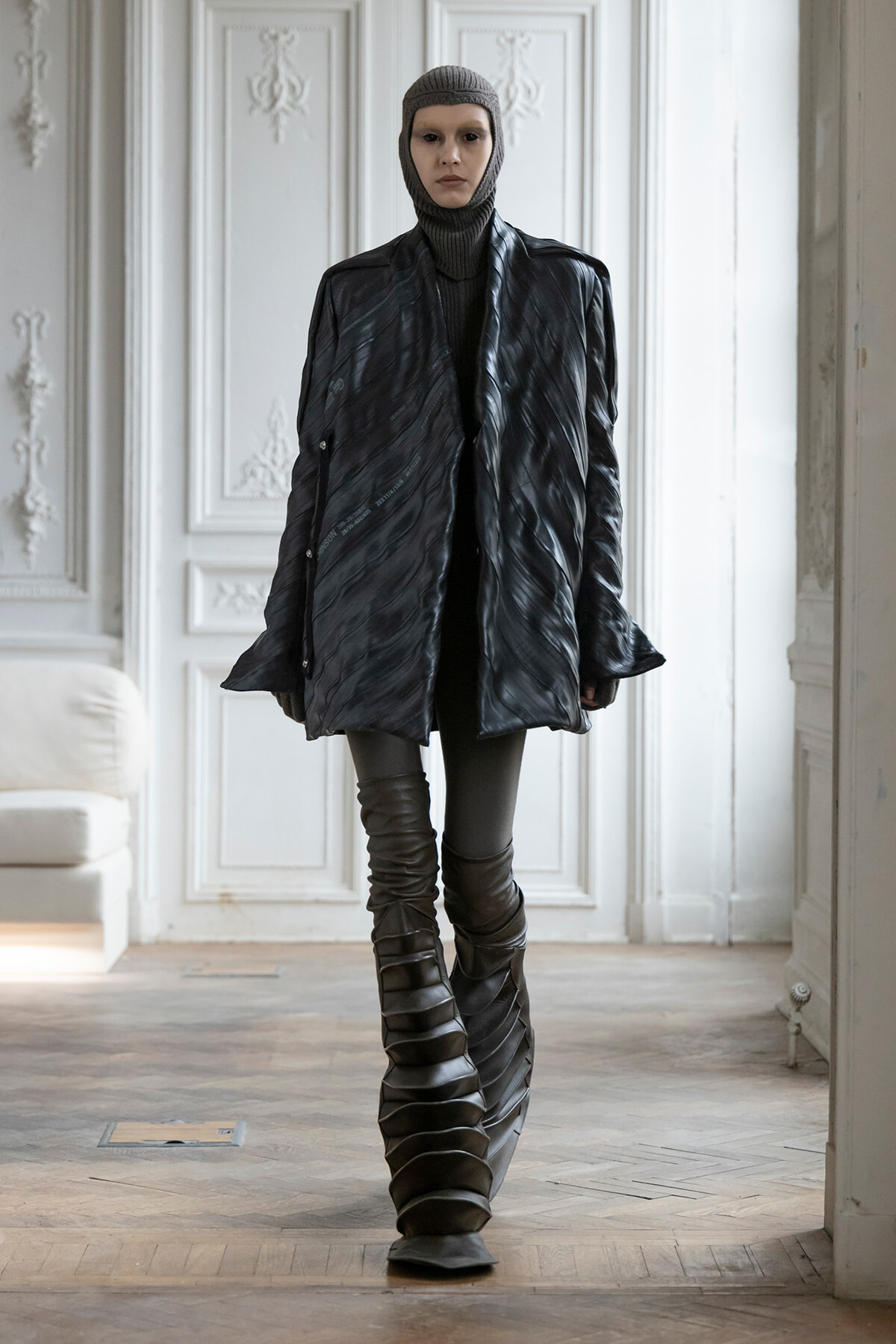 Model wearing an oversized black quilted coat, grey knit balaclava, and dramatic stacked-strap boots in a bright, ornate white room.