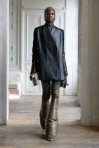 Person wearing an oversized black leather coat, gray knit balaclava and turtleneck, and tall brown boots indoors.
