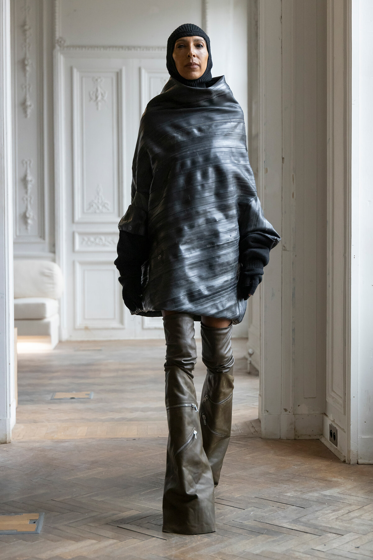 Person in a black, oversized glossy poncho with a knit hood and tall boots stands in a white, ornate room in a fashion pose.