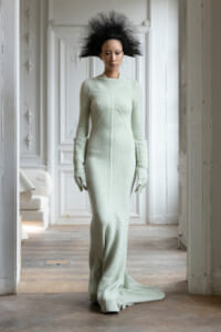 Model in a pale mint green knit gown with matching gloves standing in a white, ornate room with decorative panels and molding.