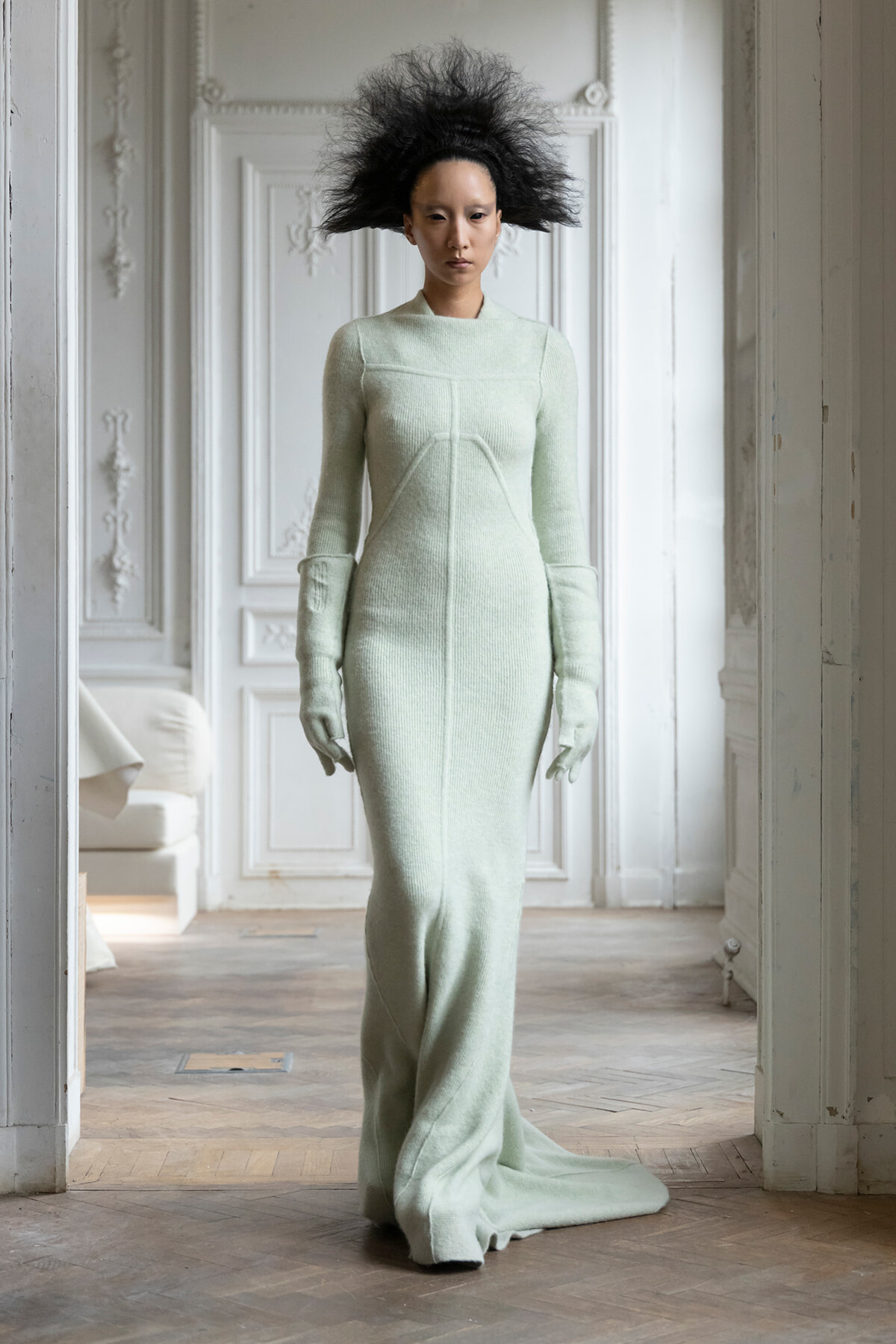 Model in a pale mint green knit gown with matching gloves standing in a white, ornate room with decorative panels and molding.