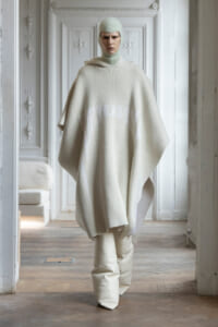 Model wearing an ivory knit poncho and matching balaclava stands in a pale, ornate room, facing the camera.