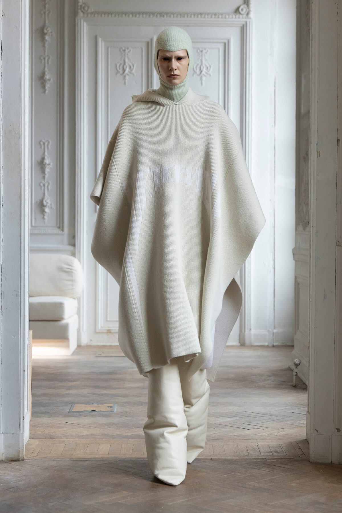 Model wearing an ivory knit poncho and matching balaclava stands in a pale, ornate room, facing the camera.