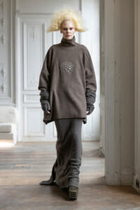 Model wearing an oversized brown fleece outfit with a circular studded design on the chest, walking in a pale ornate room.