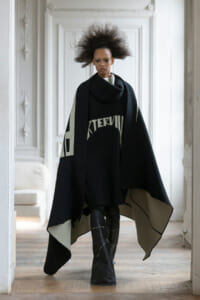 Model walking toward camera in a stark black cape with gray inner lining and bold graphic, tall boots, in a bright, ornate white room.