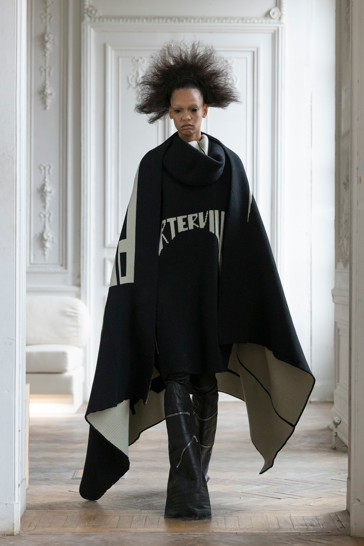 Model walking toward camera in a stark black cape with gray inner lining and bold graphic, tall boots, in a bright, ornate white room.