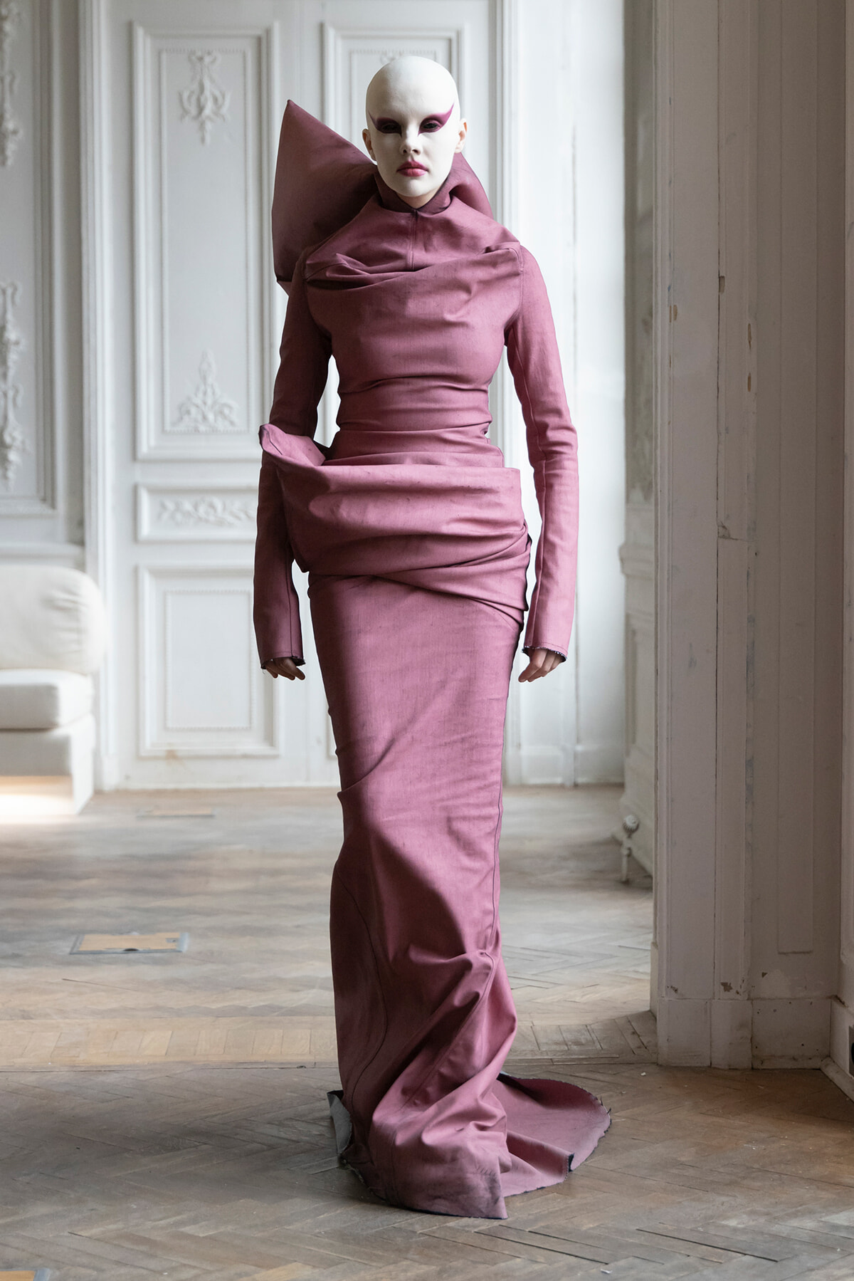Model in a dusty pink floor-length gown with an oversized shoulder collar and draped folds, wearing a white mask and pink eye makeup in a pale, ornate room.