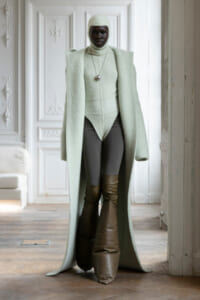 Person wearing a mint-green long coat and matching knit bodysuit with a hood, standing in a white ornate room.