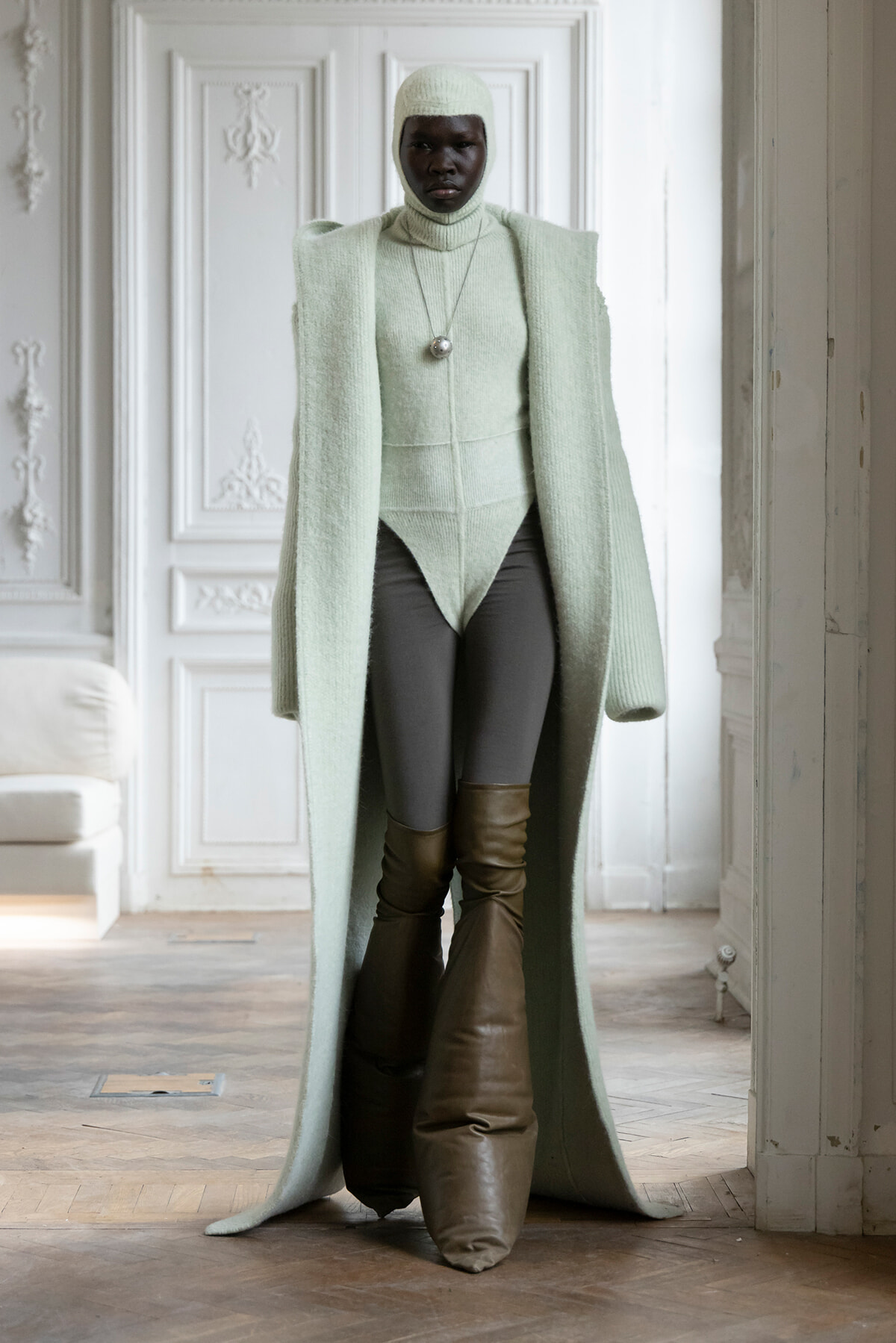 Person wearing a mint-green long coat and matching knit bodysuit with a hood, standing in a white ornate room.