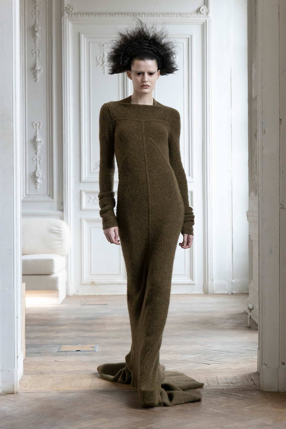 Model wearing a brown full-length knitted dress with a long train, standing in an ornate white room with dramatic black hair.