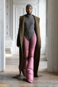 Model in a gray knit bodysuit with a matching hood, a long brown coat, and pink puffer pants indoors.
