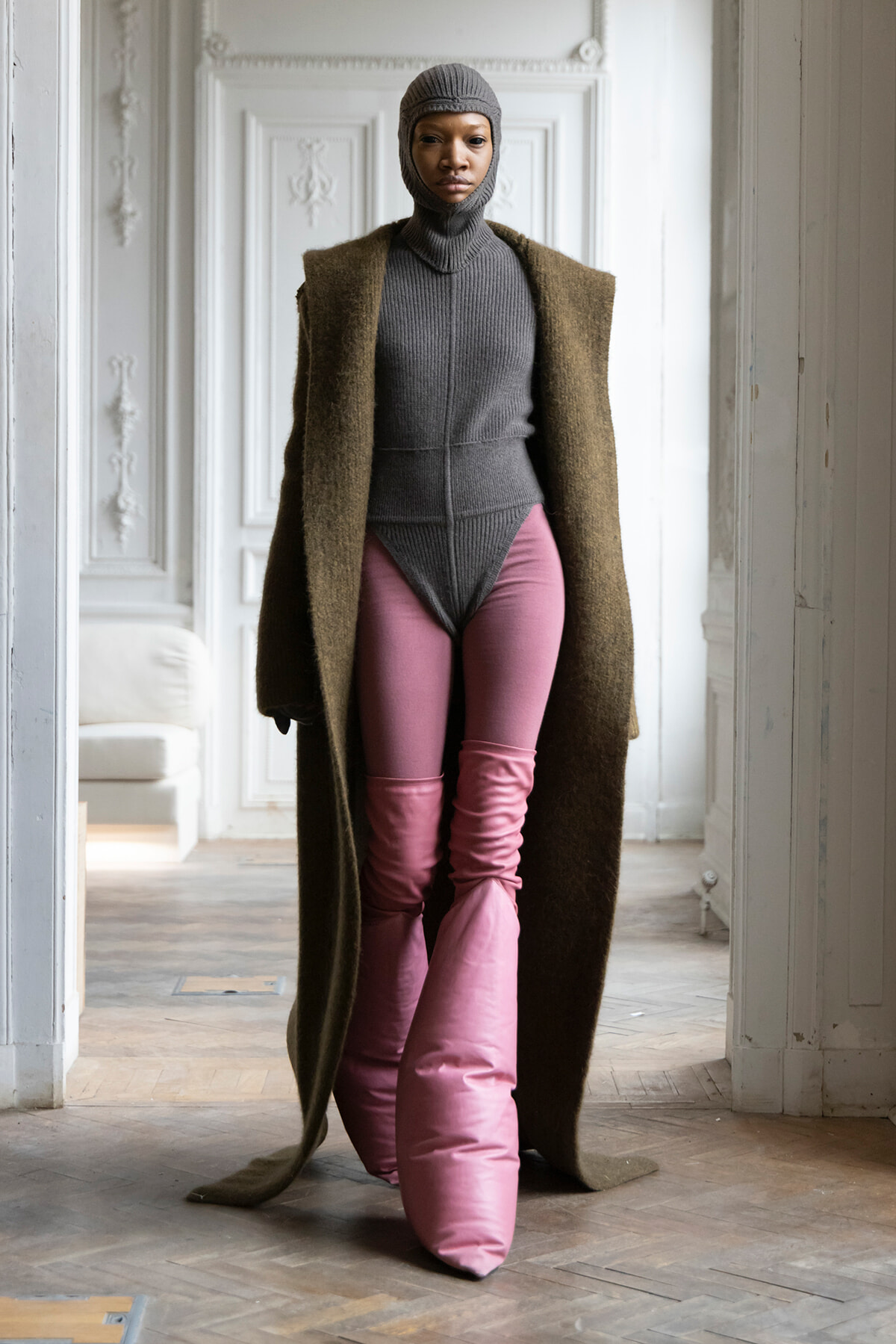 Model in a gray knit bodysuit with a matching hood, a long brown coat, and pink puffer pants indoors.
