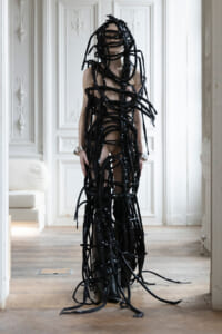 Person wearing an avant-garde black latex bondage outfit made of long strips, standing in a pale ornate room.