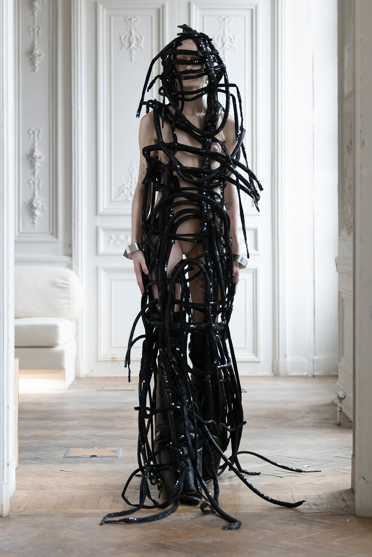 Person wearing an avant-garde black latex bondage outfit made of long strips, standing in a pale ornate room.