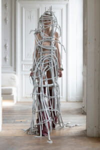 Person wrapped in grey, rope-like bands forming a sculptural bodysuit in a bright, ornate room.