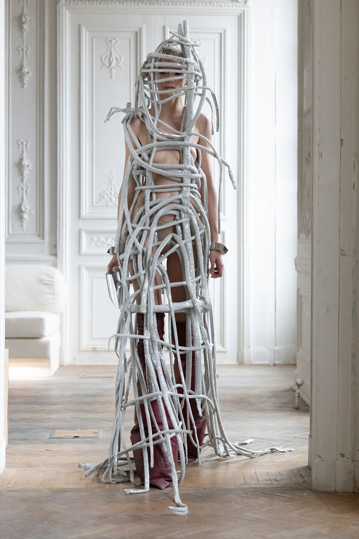 Person wrapped in grey, rope-like bands forming a sculptural bodysuit in a bright, ornate room.