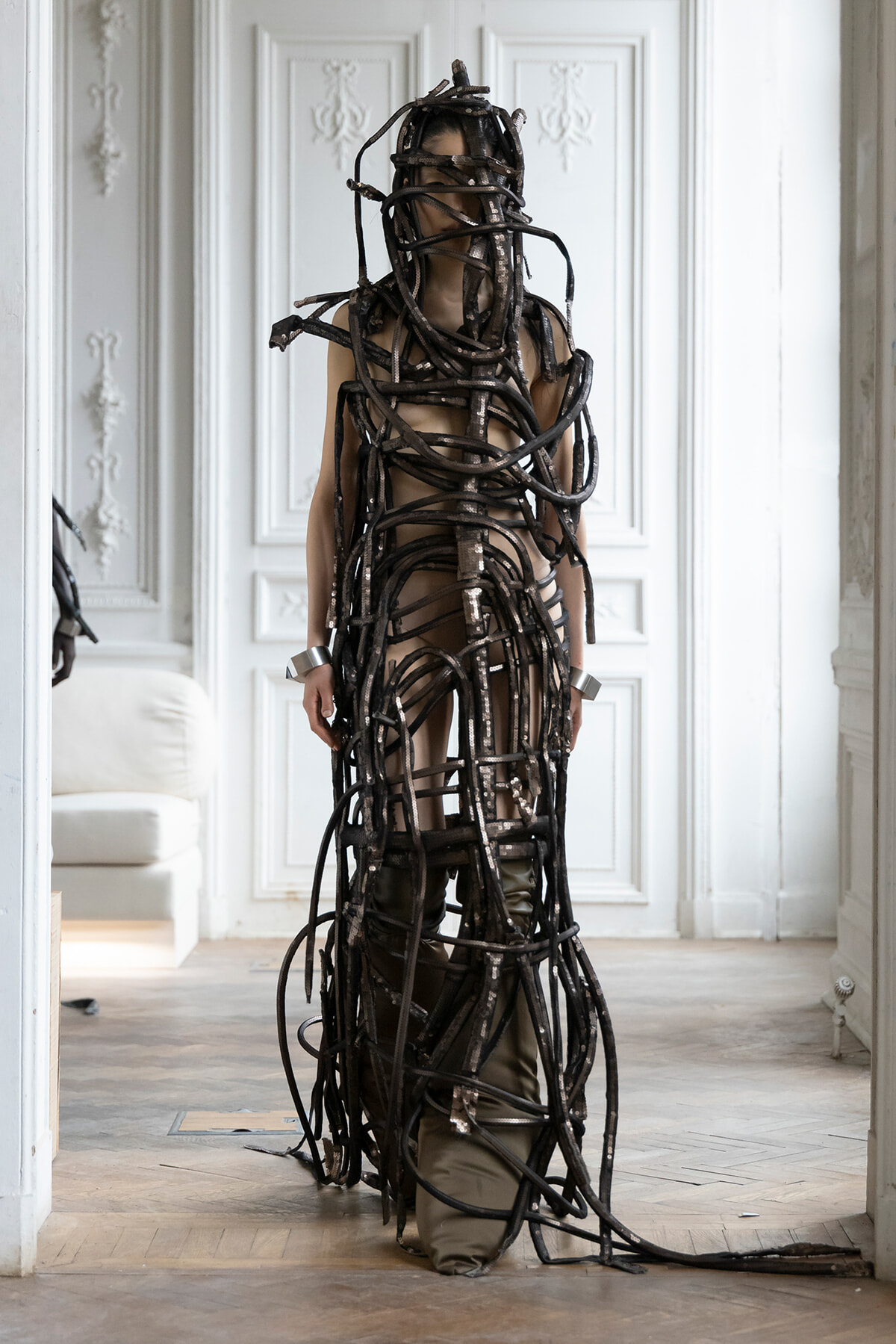 Person wrapped in a dense web of black cords covering the body and head in a white ornate room.