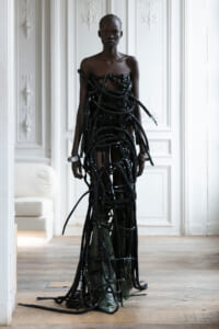 Model wearing a sculptural black rope dress made of tangled hoses, standing in a white paneled room.