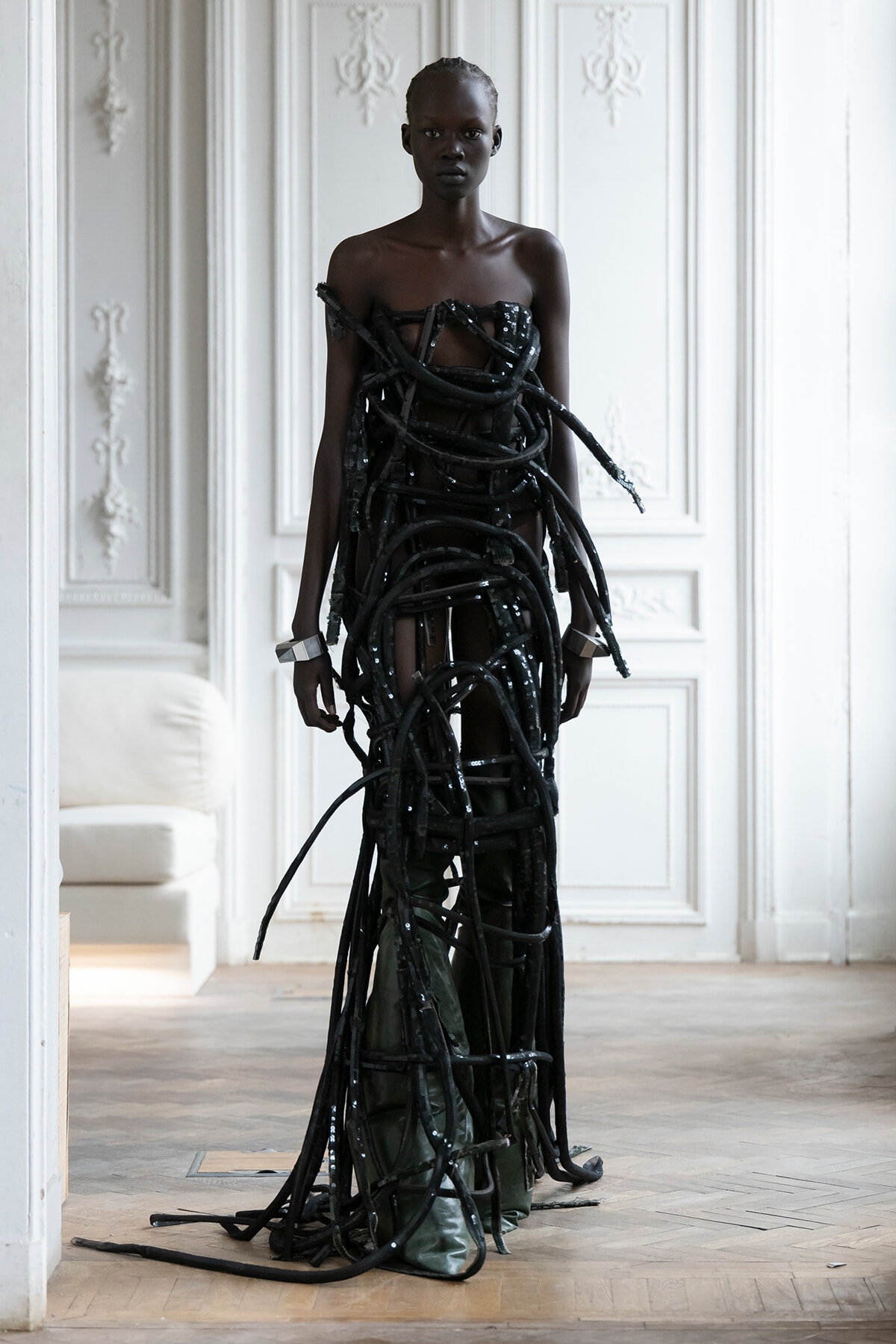 Model wearing a sculptural black rope dress made of tangled hoses, standing in a white paneled room.