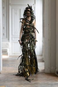 Person standing in a bright, ornate room wearing a full-body sculpture made of black, glittery straps wrapped around the torso and limbs.