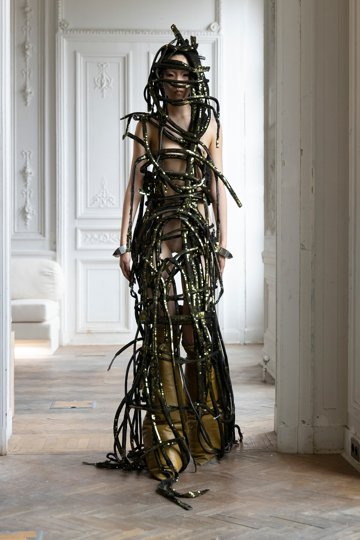 Person standing in a bright, ornate room wearing a full-body sculpture made of black, glittery straps wrapped around the torso and limbs.