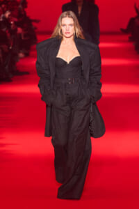 Model walking a runway in a black tailored suit with a bustier top, under red lighting.