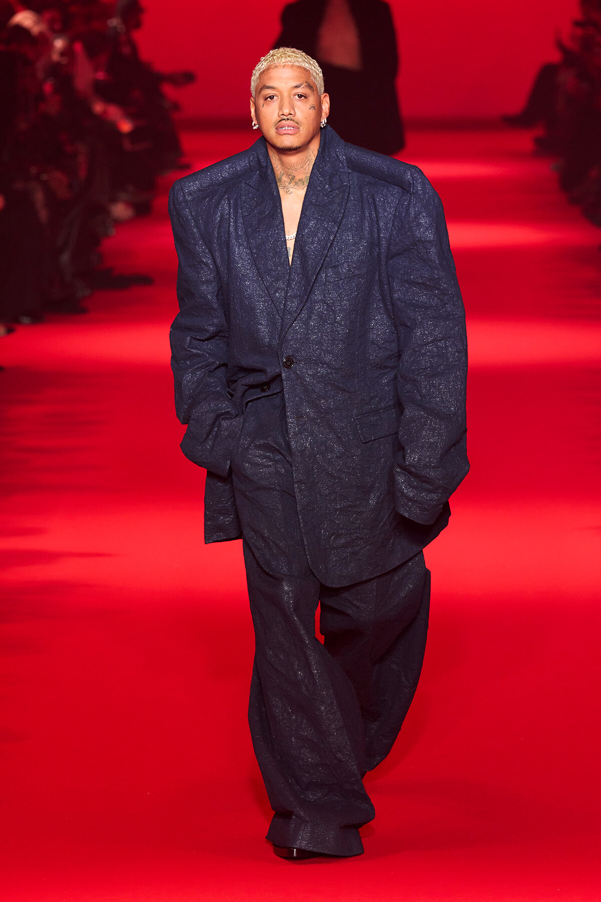 Male model with bleached blond hair wearing an oversized dark blue textured suit, walking down a red-lit fashion show runway.