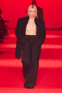 Model walks the runway with an oversized black blazer open over a bare chest on a red-lit catwalk.