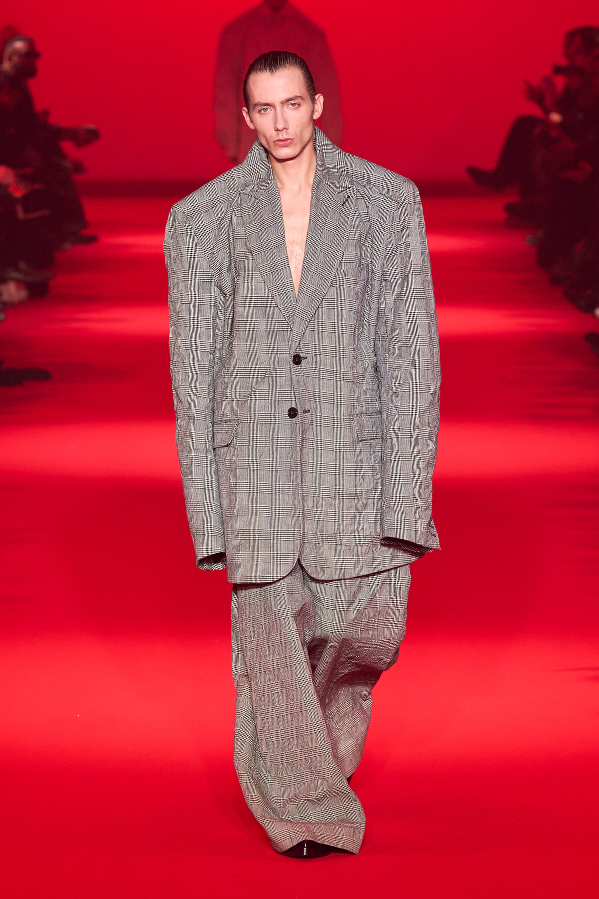 Male model in an oversized gray plaid suit walks a red runway at a fashion show.