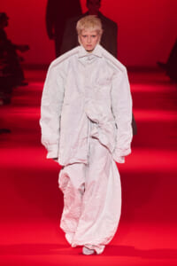 Model walks the runway in an oversized wrinkled white coat and matching pants, against a red-lit backdrop.