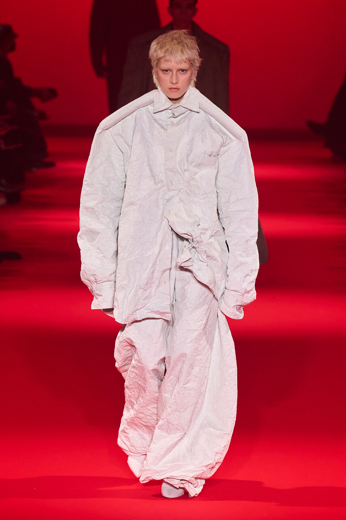 Model walks the runway in an oversized wrinkled white coat and matching pants, against a red-lit backdrop.
